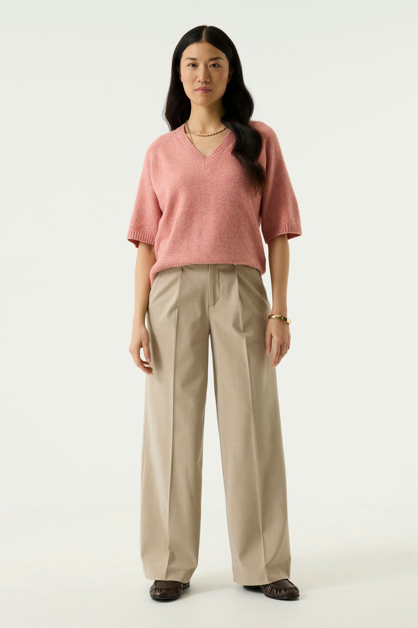 Woman modelling the Nora V-Neck Knit in pink with wide-leg beige trousers and loafers, full-length view.