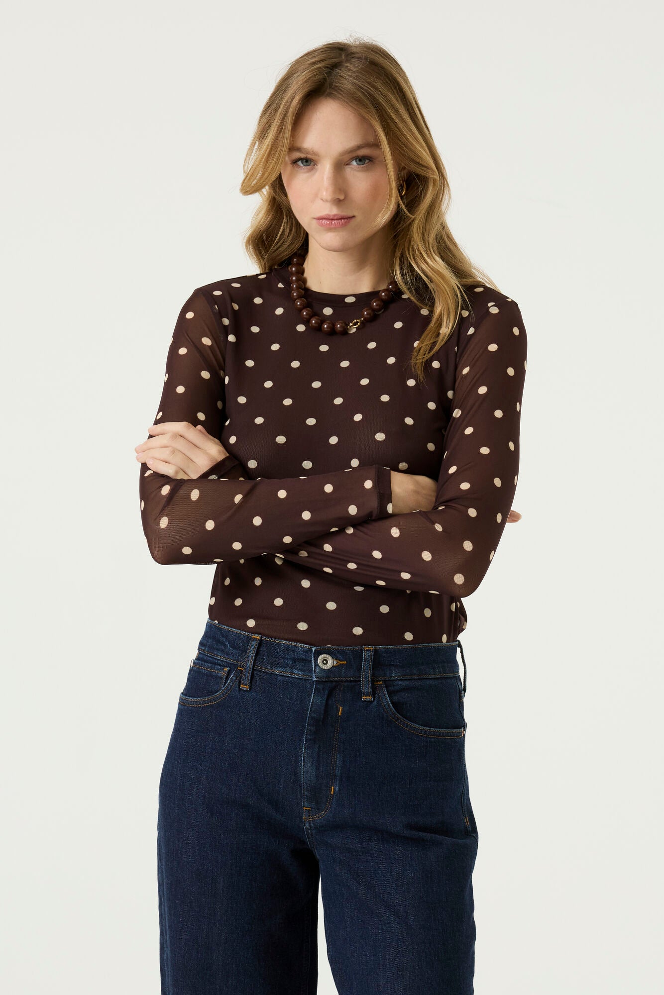 Woman wearing a brown long-sleeve mesh top with cream polka dots, dark blue jeans