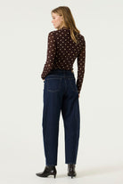 Woman wearing a brown long-sleeve mesh top with cream polka dots, dark blue jeans, and black heeled ankle boots.