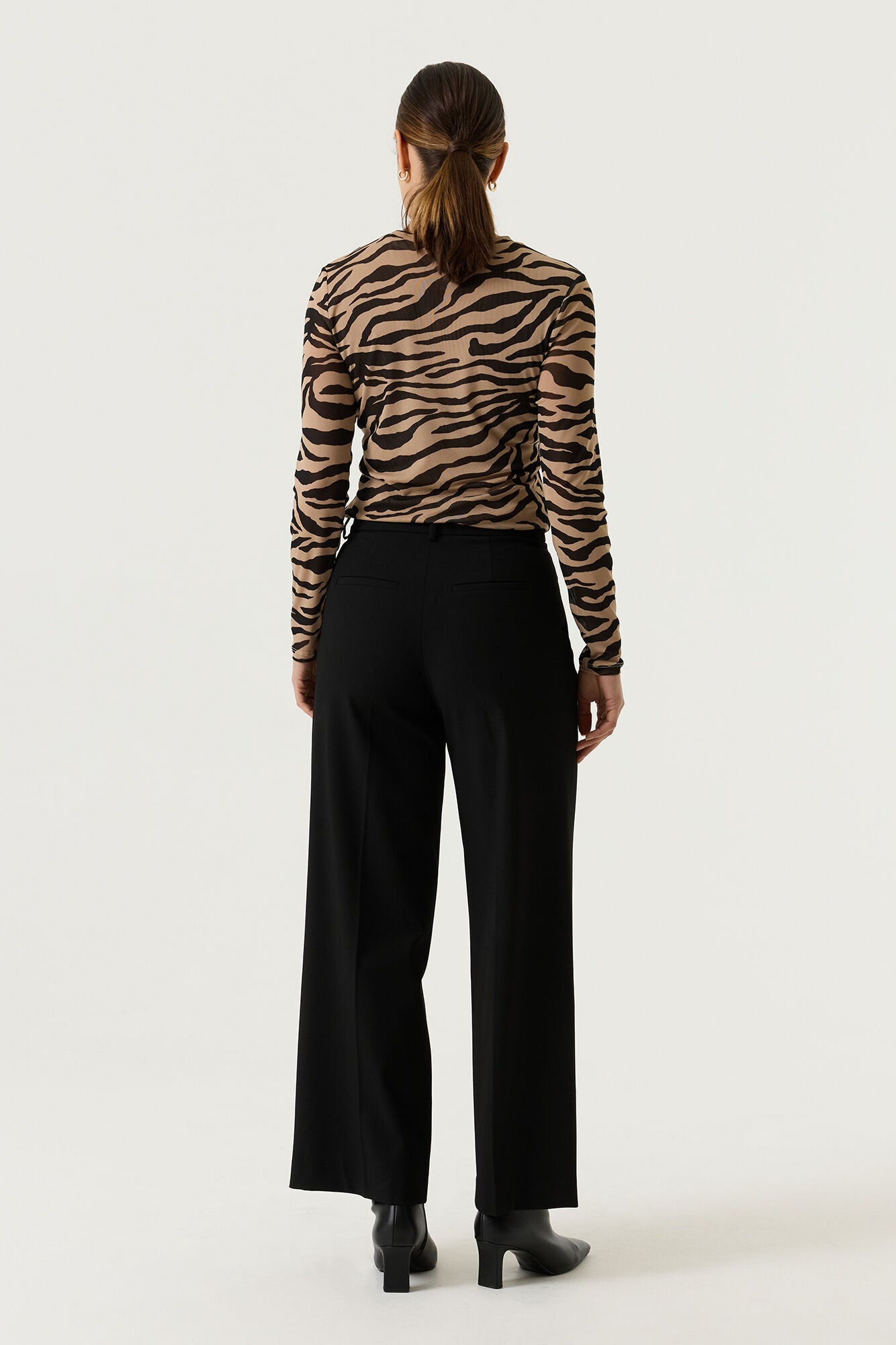 Back view of a woman wearing a tan and black zebra-print long-sleeve top with black wide-leg trousers and black heeled ankle boots.