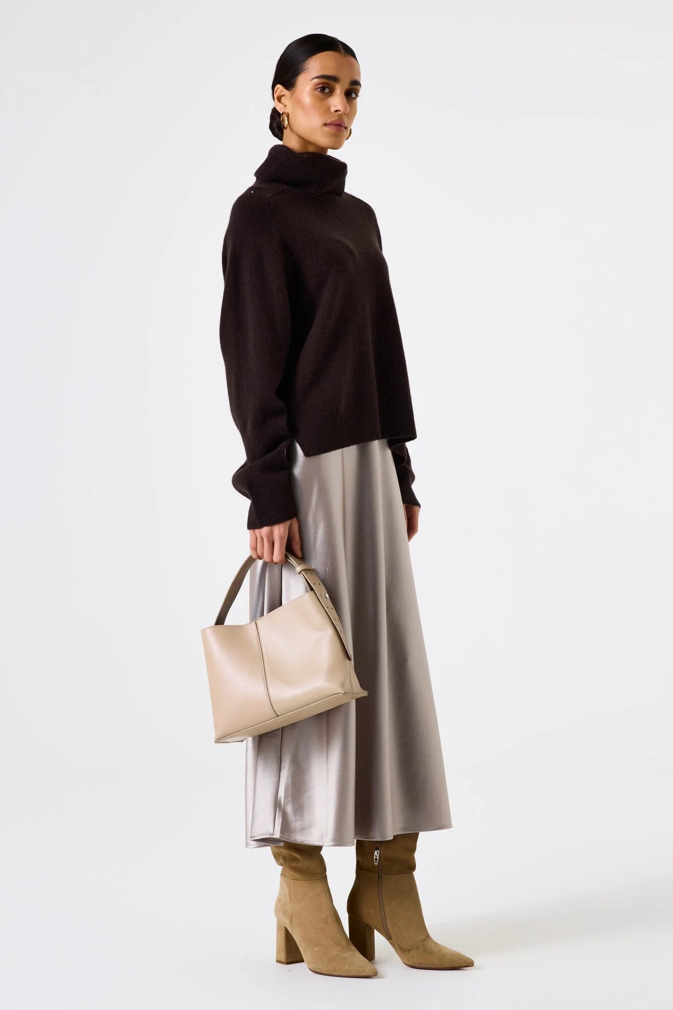Woman wearing a brown turtleneck, silver skirt, and beige boots, holding a beige handbag on a white background.