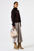 Woman wearing a brown turtleneck, silver skirt, and beige boots, holding a beige handbag on a white background.