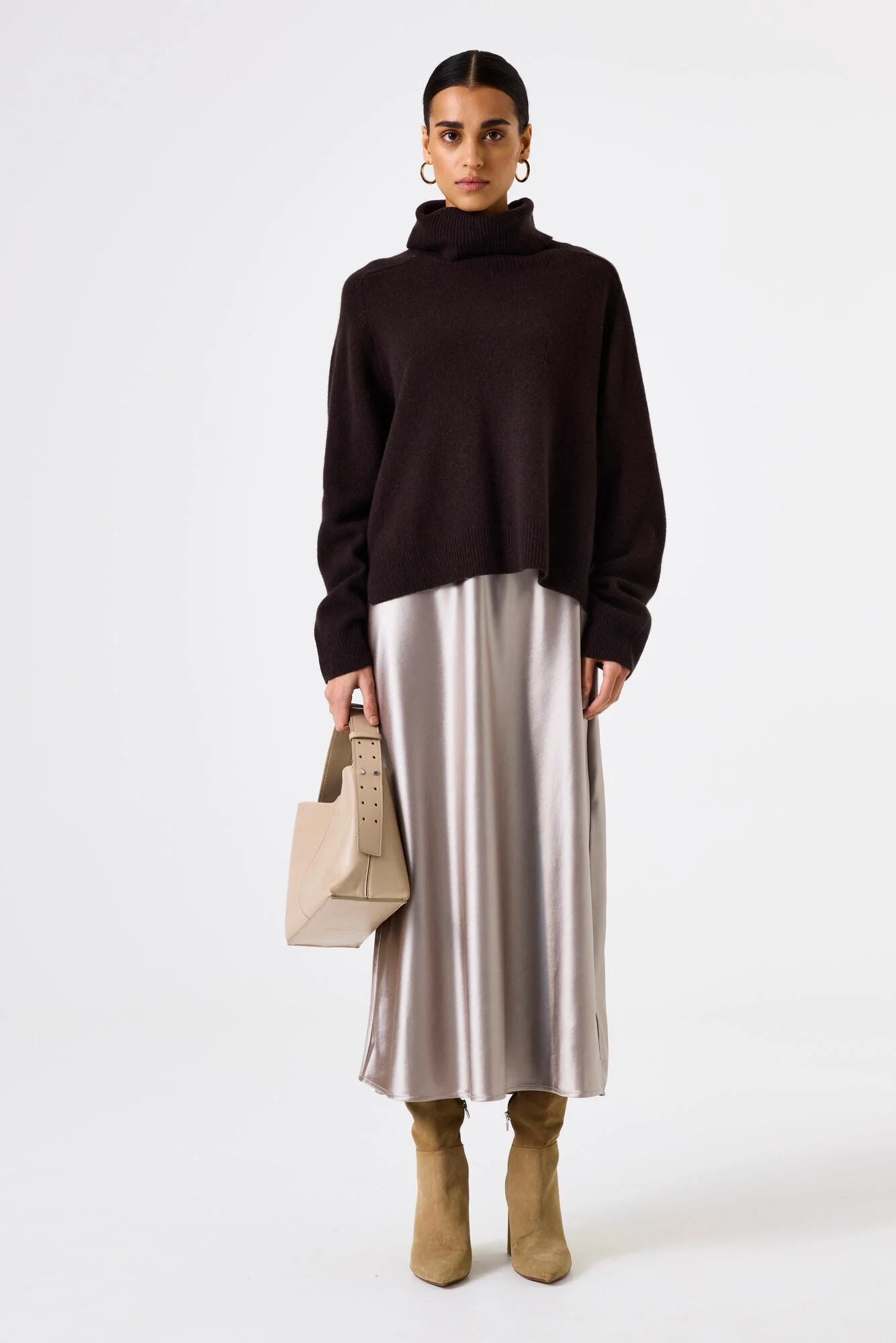 Woman wearing a dark brown sweater and silver skirt with beige boots, holding a beige handbag on a white background.