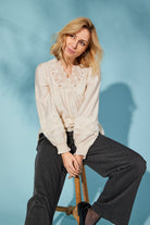 Woman sitting on a stool wearing a cream blouse and dark pants against a light blue background
