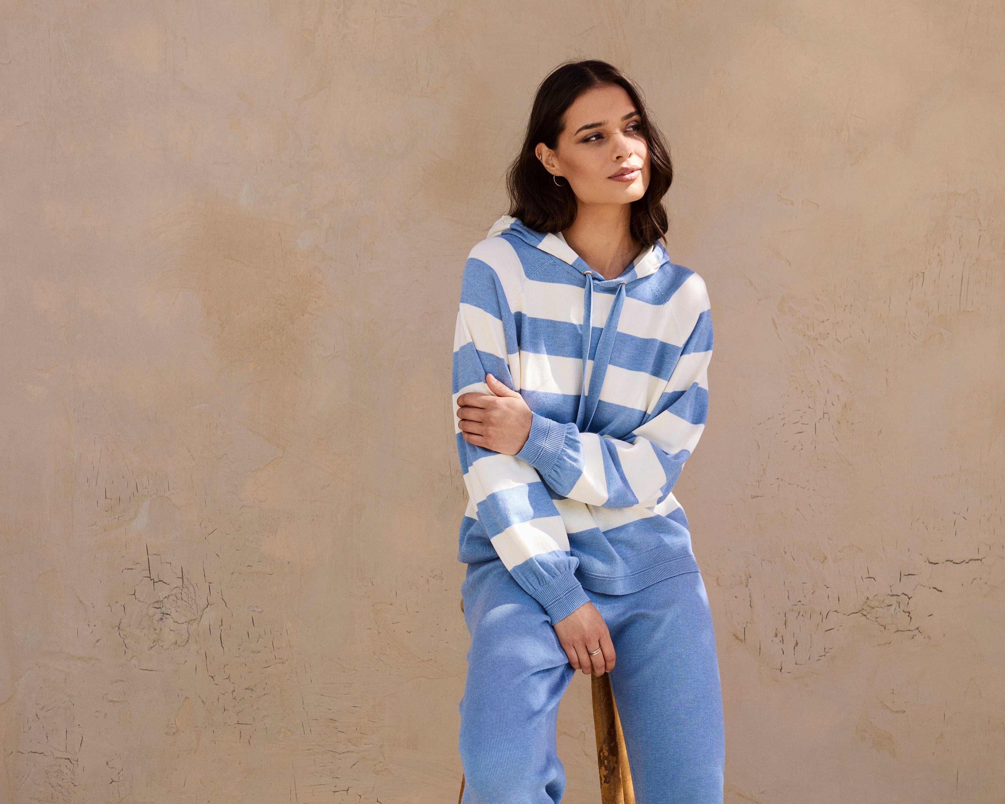 Woman wearing a blue and white striped sweater and pants sitting on a wooden stool against a beige wall.