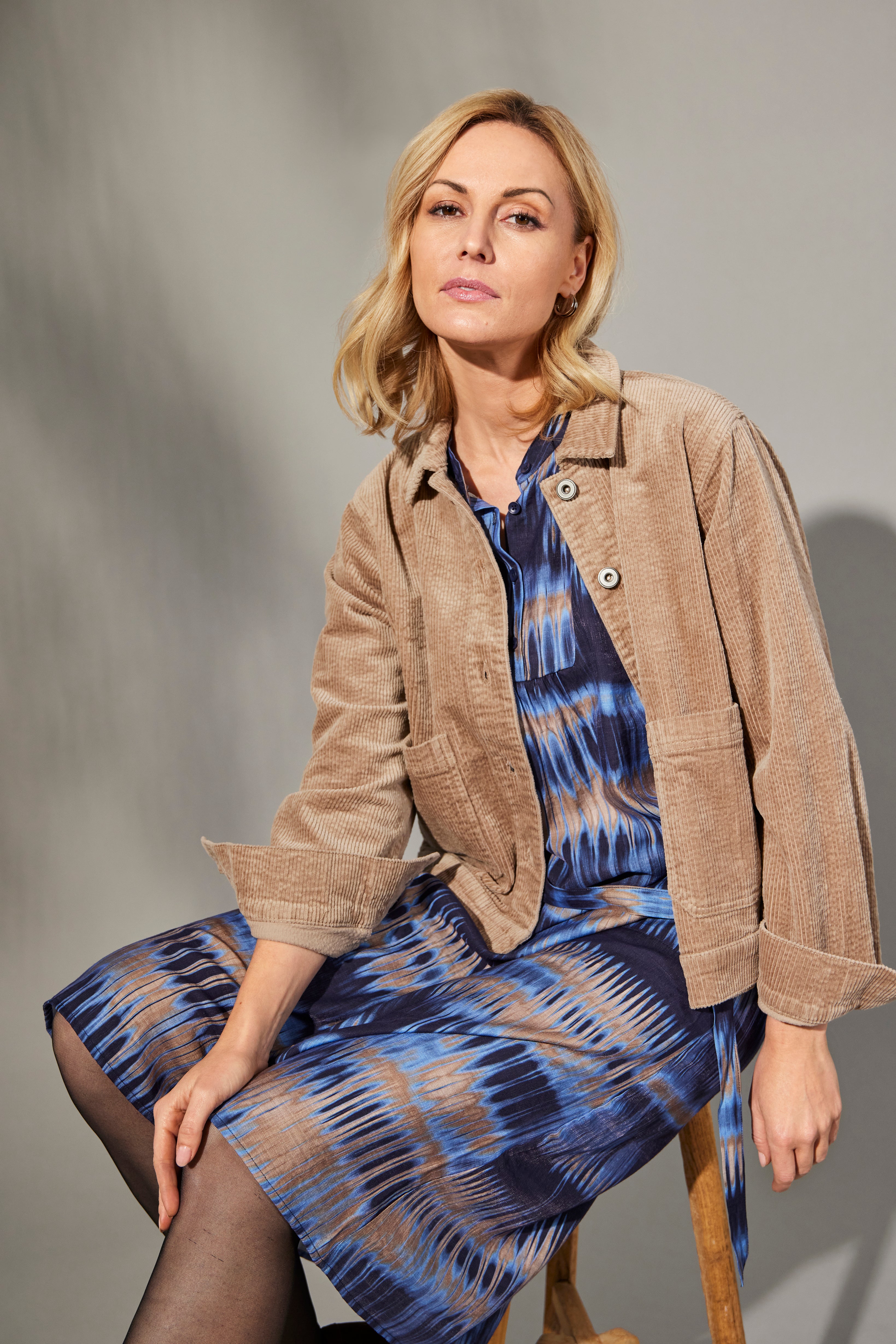 Woman wearing a beige jacket over a blue patterned dress on a gray background