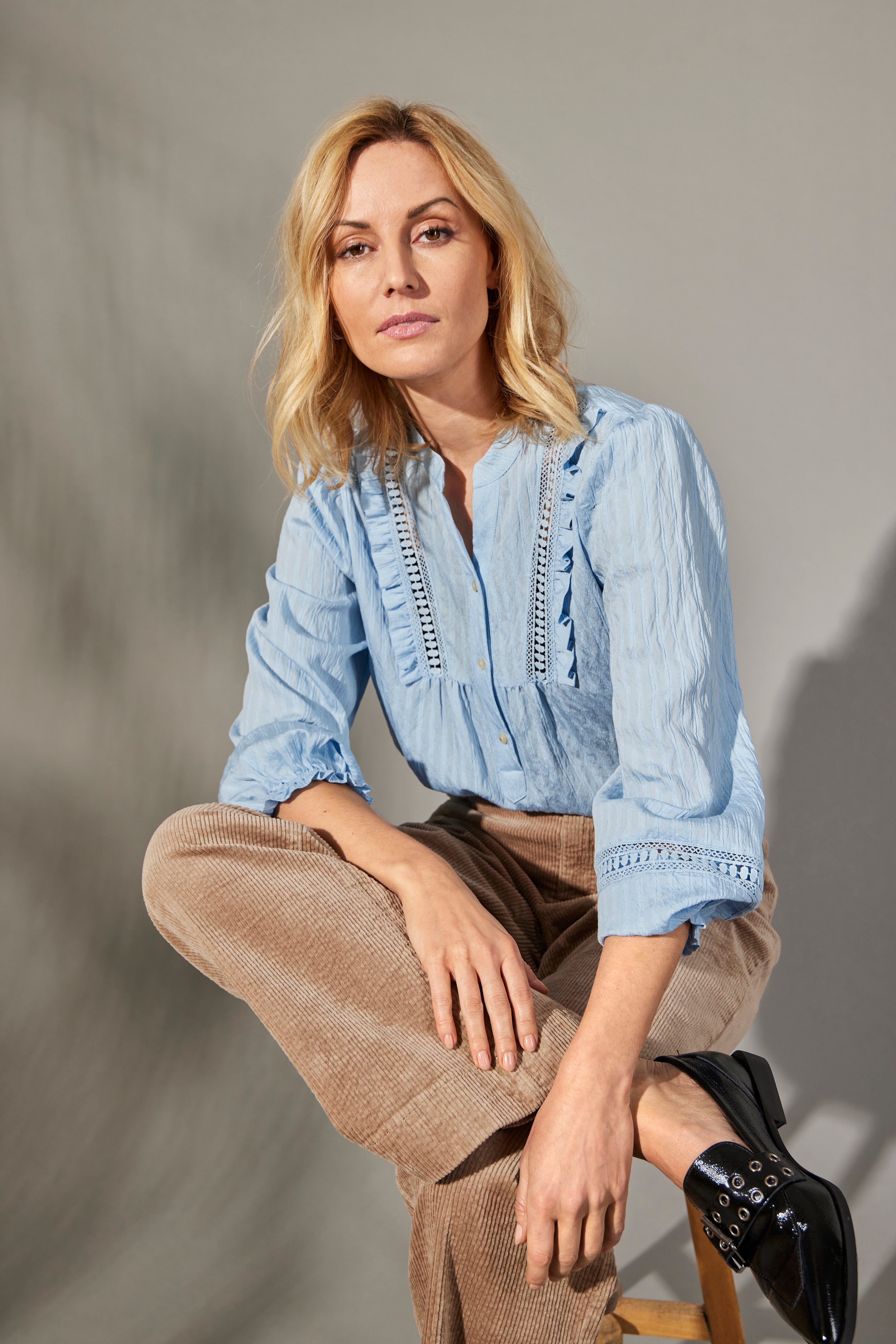 Woman wearing a blue blouse with intricate details and beige pants sitting on a stool against a plain background.